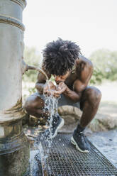 Young man during workout, drinking from fountain in a park - FBAF01239