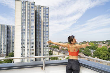 Rückenansicht einer glücklichen Frau, die mit ausgestreckten Armen auf einer Dachterrasse steht - FBAF01246