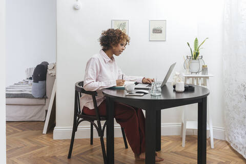 Junge Frau, die zu Hause arbeitet, Notizen macht und einen Laptop benutzt, lizenzfreies Stockfoto