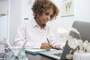 Junge Frau, die zu Hause arbeitet, Notizen macht und einen Laptop benutzt - VPIF01954