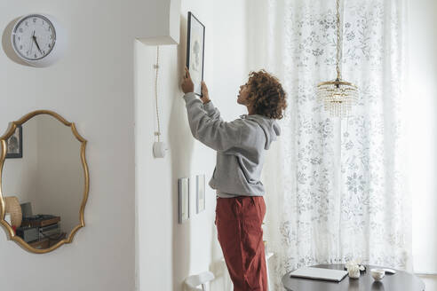 Young woman hanging up picture at home - VPIF01973