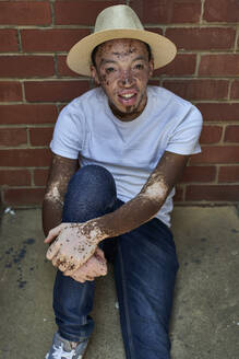 Portrait of young man with vitiligo wearing a hat, sitting on the floor - VEGF01328