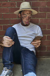 Portrait of young man with vitiligo wearing a hat, checking his phone sitting on the floor - VEGF01329