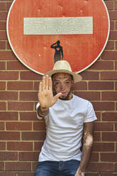 Portrait of young man with vitiligo wearing a hat doing a stop sign with his hand on a forbidden sign - VEGF01331