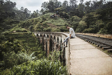 Sri Lanka, Uva Province, Demodara, Portrait of young man sitting at edge of Nine Arch Bridge - DAWF01142