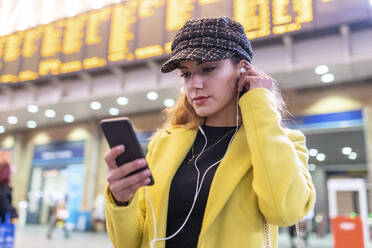 Frau am Bahnhof, die ihr Smartphone überprüft und Kopfhörer benutzt - WPEF02435