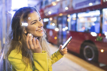 Glückliche Frau, die auf den Bus wartet und Musik hört, in einer nächtlichen Stadt, London, Großbritannien - WPEF02442