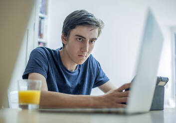 Konzentrierter Jugendlicher mit Laptop und Tablet auf dem Tisch zu Hause - AJOF00083