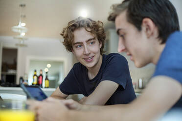 Lächelnder junger Mann sieht seinen Freund an, der zu Hause sein Smartphone benutzt - AJOF00088
