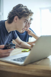 Foused teenage boy using laptop and smartphone on table at home - AJOF00089