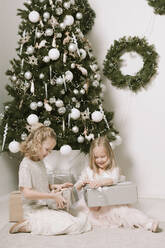 Zwei kleine Mädchen sitzen vor dem Weihnachtsbaum und öffnen Weihnachtsgeschenke - EYAF00802