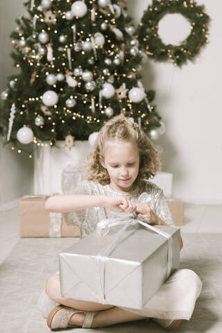 Porträt eines blonden kleinen Mädchens, das vor einem Weihnachtsbaum sitzt und ein Weihnachtsgeschenk öffnet, lizenzfreies Stockfoto