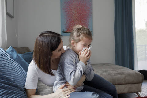 Mother sitting with daughter blowing her nose on couch in living room at home stock photo