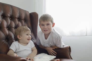 Portrait of boy with his baby brother in leather armchair in living room at home - KMKF01160