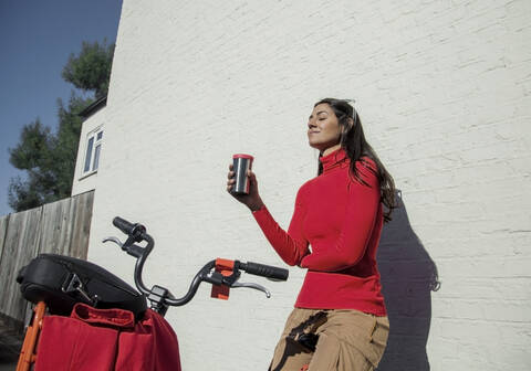 Junge Frau mit Fahrrad, an die Wand gelehnt, trinkt Kaffee aus einem wiederverwendbaren Becher, lizenzfreies Stockfoto