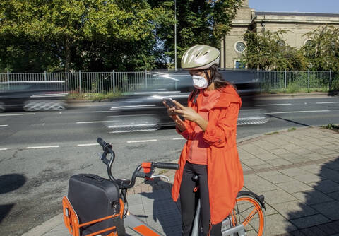Junge Frau mit Helm und Gesichtsmaske, auf dem Fahrrad sitzend, mit Smartphone, lizenzfreies Stockfoto