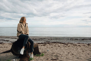 Fröhliche junge Frau sitzt auf verwittertem Metall am Strand gegen den Himmel - MASF16082