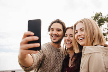 Lächelnder Mann nimmt Selfie mit Freunden auf Smartphone gegen Himmel - MASF16087