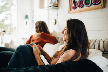 Lächelnde Frau mit Smartphone, die wegschaut, während sie mit einem Freund auf dem Sofa zu Hause sitzt - MASF16104