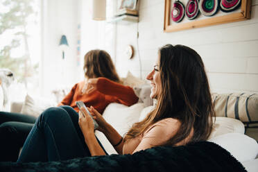 Smiling woman with smart phone looking away while sitting with friend on sofa at home - MASF16104