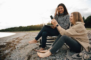 Happy friends looking at smart phone while sitting on shore at beach against sky - MASF16114