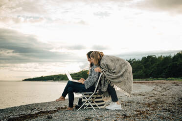 Friend greeting woman working on laptop while sitting at beach against sky - MASF16117