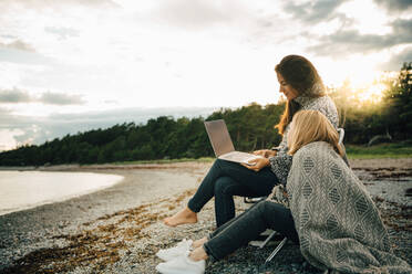 Woman wrapped in blanket looking at friend using laptop on sea shore - MASF16118