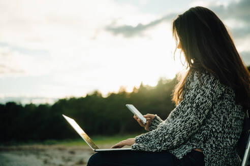 Mittlere erwachsene Frau mit Laptop, die bei Sonnenuntergang am Strand sitzt und Textnachrichten schreibt - MASF16119