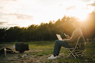 Nachdenklicher Mann mit Laptop und Handy auf einem Stuhl sitzend bei Sonnenuntergang in voller Länge - MASF16124