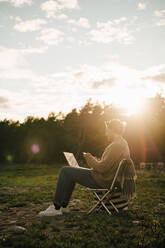 Full length of man with laptop using mobile phone while sitting on chair during sunset - MASF16125