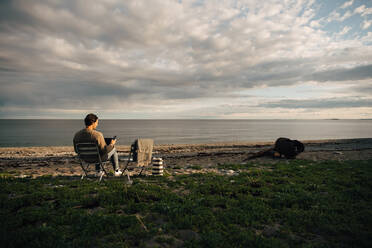 Mann in voller Länge, der Laptop und Mobiltelefon benutzt, während er am Strand vor einem bewölkten Himmel sitzt - MASF16126