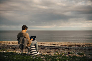 Mid adult man using laptop and mobile phone while sitting on sea shore at beach against sky - MASF16127