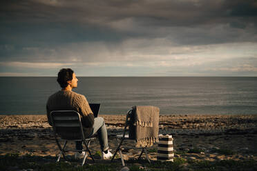 Nachdenklicher Mann, der einen Laptop benutzt, während er am Strand vor einem bewölkten Himmel sitzt - MASF16128