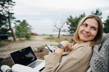 Lächelnde junge Frau mit Technologien, die wegschauen, während sie im Urlaub auf einem Stuhl sitzt - MASF16136