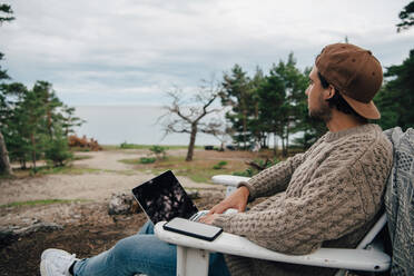 Nachdenklicher erwachsener Mann mit Laptop und Mobiltelefon auf einem Stuhl sitzend im Urlaub - MASF16140