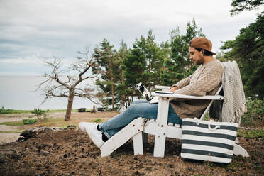 Side view of man using laptop while sitting on Adirondack chair at forest - MASF16143