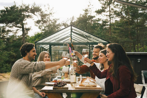 Männliche und weibliche Freunde stoßen mit Weingläsern an, während sie das Abendessen auf einer Gartenparty genießen, lizenzfreies Stockfoto