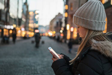 Teenager-Mädchen mit Strickmütze Textnachrichten auf dem Smartphone in der Stadt bei Sonnenuntergang - MASF16156