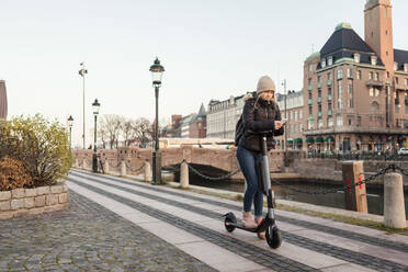 Teenager-Mädchen Text-Messaging auf dem Smartphone während der Fahrt E-Scooter auf der Straße am Kanal in der Stadt gegen den Himmel - MASF16161