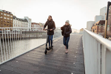 Glückliche weibliche Freunde genießen mit elektrischen Push-Scooter auf Brücke in der Stadt - MASF16167