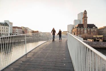 Teenager-Mädchen zu Fuß mit Freund fahren Push-Scooter auf Brücke in der Stadt gegen klaren Himmel - MASF16168