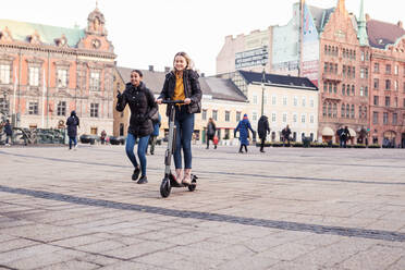 Friend walking behind teenage girl riding e-scooter on street in city - MASF16170