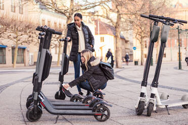 Teenager unlocking electric push scooter with smart phone while crouching by friend on street in city - MASF16171