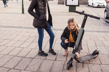 Teenager-Mädchen scannt Handy über elektrischen Roller, während sie bei einem Freund auf der Straße hockt - MASF16172