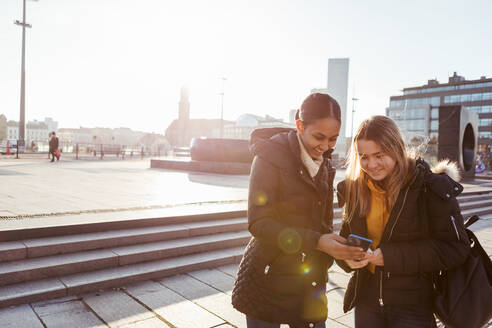 Lächelnde Teenager-Freunde, die auf ihr Smartphone schauen, während sie an einem sonnigen Tag in der Stadt stehen - MASF16188