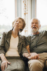 Senior couple holding hands while sitting against window at nursing home - MASF16201