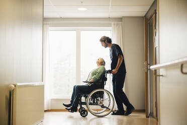 Vollständige Seitenansicht eines Pflegers, der eine behinderte ältere Frau im Rollstuhl in einer Gasse eines Altenheims schiebt - MASF16206