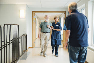 Ein älterer Mann geht Arm in Arm mit einer Krankenschwester, während er einen Freund in einer Gasse eines Pflegeheims betrachtet - MASF16217
