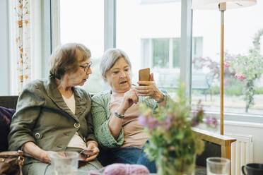 Ältere Frau, die ein Mobiltelefon benutzt, während sie mit einer Freundin auf dem Sofa vor dem Fenster eines Altersheims sitzt - MASF16220