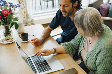 Confident male caregiver assisting senior woman doing online shopping with credit card and laptop in nursing home - MASF16232